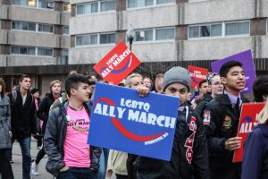 Students march down High Avenue chanting "1, 2, 3, 4 love is what we're fighting for" while carrying signs. 