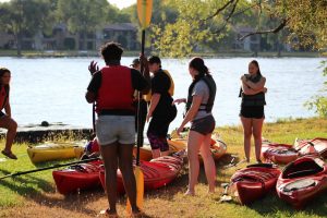 A group of students stand next to their kayaks as they prepare to get in the water.