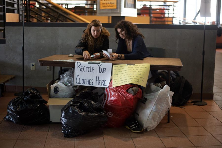 Two students sit a table with bags of clothing under the table